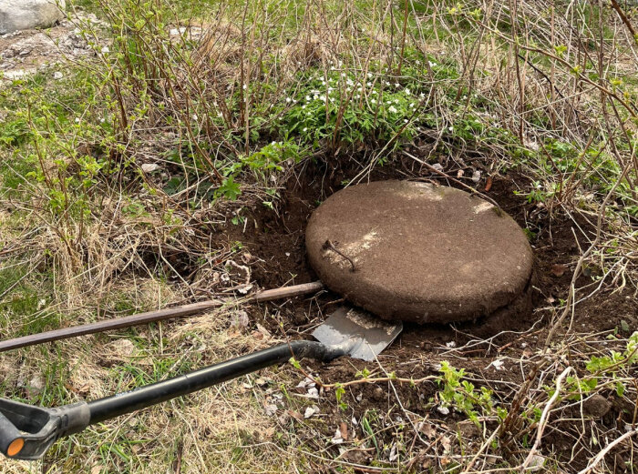 Ett betonglock i marken omgivet av vegetation och verktyg, trolig ingång till en gammal brunn.