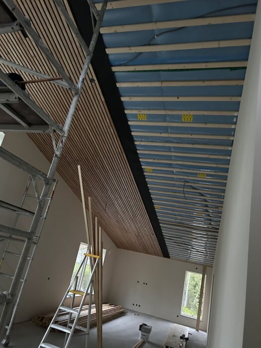 Installation av hemgjord akustikpanel med betsade fururibbor på taket och byggnadsställning i rum under renovering.
