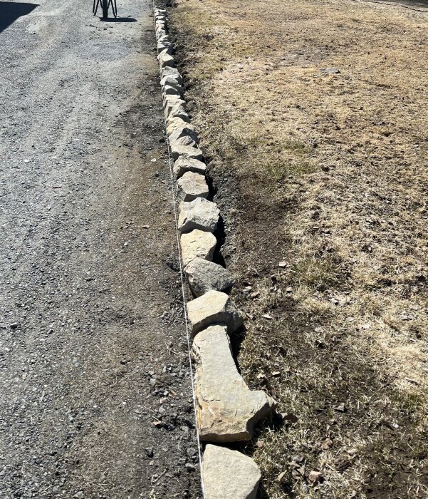 Nyupplagd kant av oregelbundna stenar mellan grusväg och gräsmatta, betong ska läggas under.