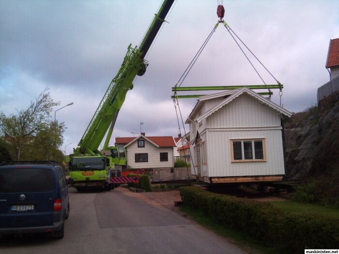 Mobilkran lyfter en vit stuga över en uppfart medan hus och fordon syns i bakgrunden.