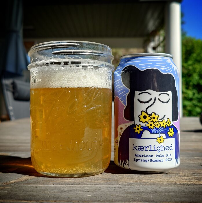 Öppnad burk av öl med etikett 'kaerlighed' bredvid ett glasburkmugg med öl på träbord utomhus.