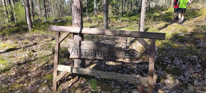 Träskylt med texten 'ÄTTESTUPAN' som pekar åt höger på en stig i skogen, med två personer som vandrar.