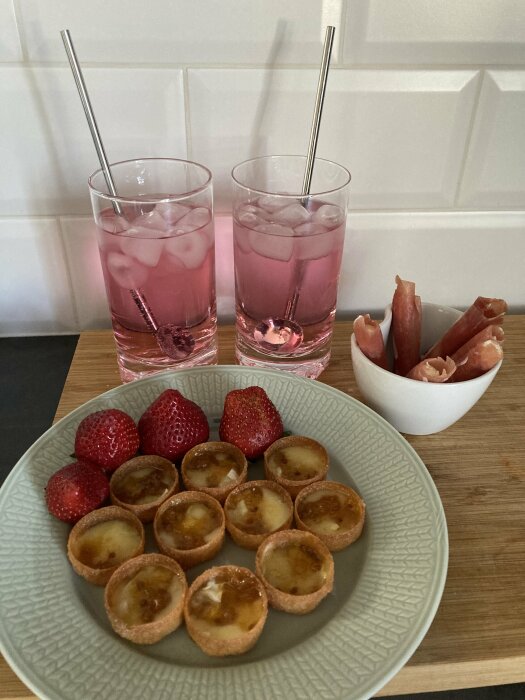 Två glas med rosa tonic och is, mini pajer samt jordgubbar och skinkrullar på ett köksbord.