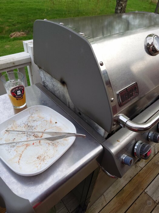 Grill med öppet lock, tom tallrik med grillspett och ett glas öl på en träterrass.