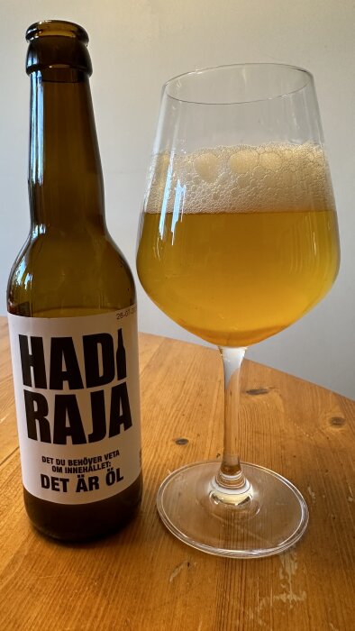 En flaska öl bredvid ett glas med öl på ett träbord med text på etiketten "HADI RAJA DET ÄR ÖL".