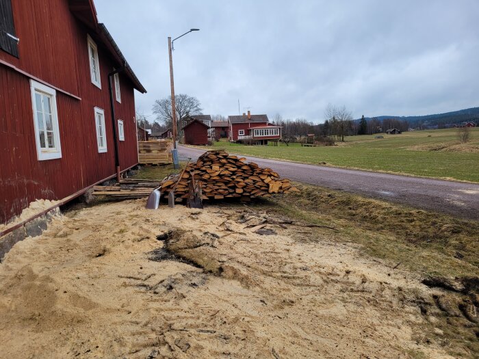 Vedstapel vid röd lantlig byggnad med sandig mark i förgrunden och gröna fält i bakgrunden.