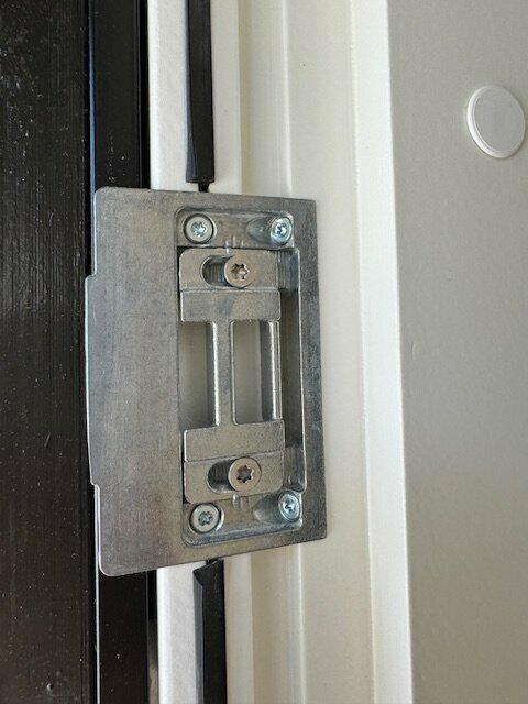 Metallfäste för dörrlås på en vit dörrkarm, ifrågasatt för eventuell borttagning eller modifiering.