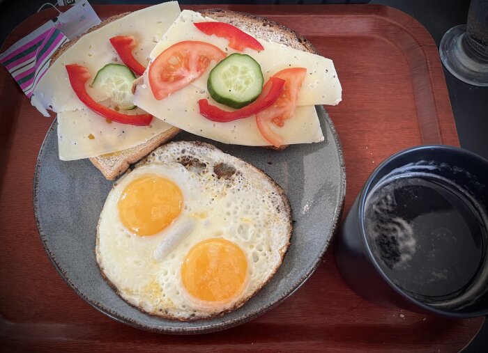 Frukosttallrik med två stekta ägg, smörgåsar med ost, tomater och gurka, och en kopp te.