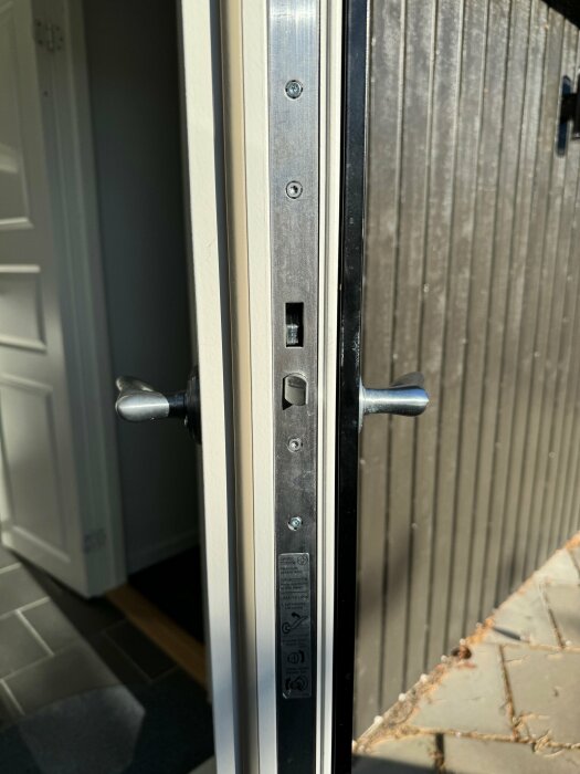 Detaljbild av en vit ytterdörrs sidoprofil som visar dörrhandtag, låskista och säkerhetsinstruktioner i soligt ljus.