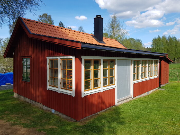 Nybyggt orangeri med rödfärgad vägg och vita fönsterpartier under blå himmel.