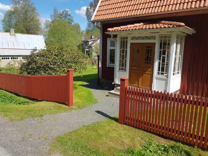Nymålat rött staket framför en röd stuga med gult dörrparti under en solig dag.