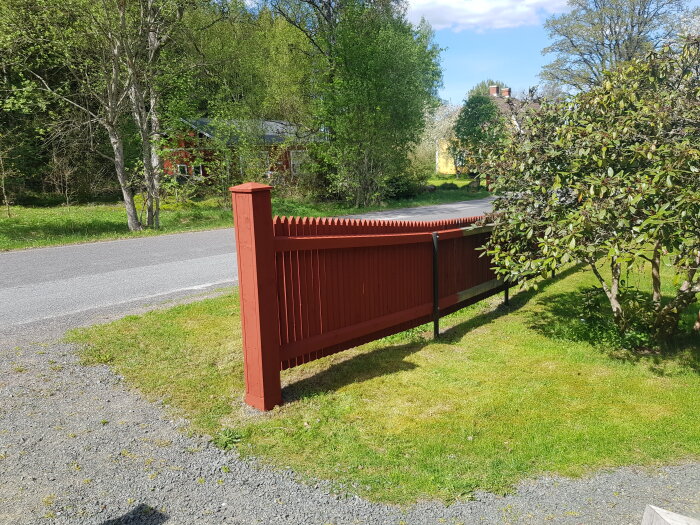 Ny målad röd trästaket vid en lummig trädgård intill en grusväg och grönska.