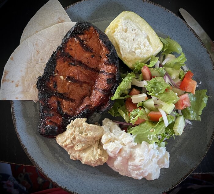 Marinerad grillad fläskkarré med sallad, potatissallad, hummus, bröd och grillad squash på tallrik.