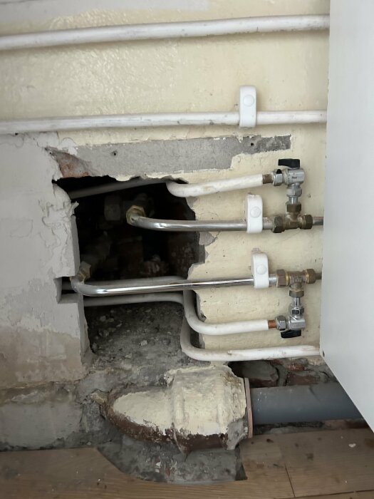 Vattenrör och avstängningsventiler bakom hål i väggen, nära golvet i ett kök.