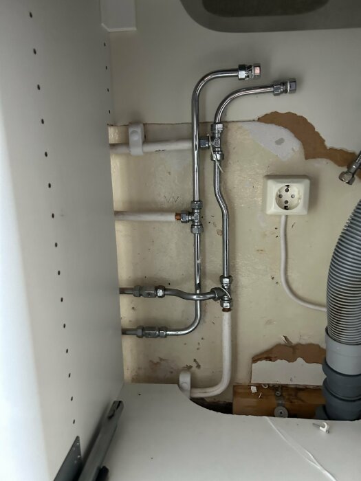 Rörinstallation bakom diskmaskin med synliga rör som går från köket till badrummet vid en sliten vägg.