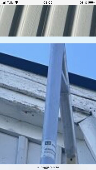 Närbild av en byggnads vitmålade träpanel med synliga sprickor och tecken på röta nära taket, vid sidan av en skylift.