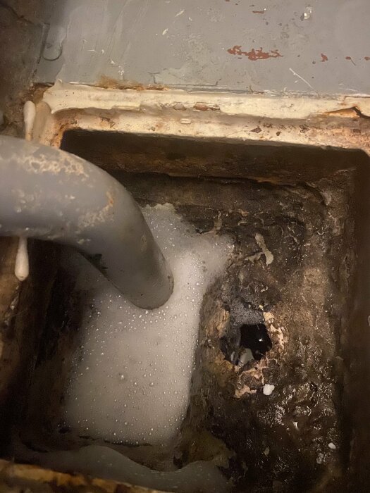 Ett skadat avloppshål med skum på golvet i en smutsig och sliten gammal toalett, indikationer på behov av underhåll.