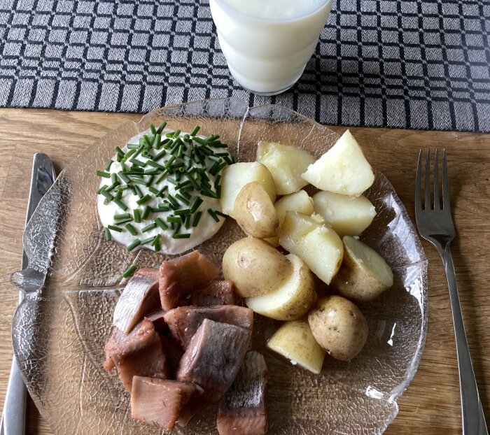 Tallrik med sill, färskpotatis och gräddfil toppad med gräslök, bredvid ett glas mjölk.