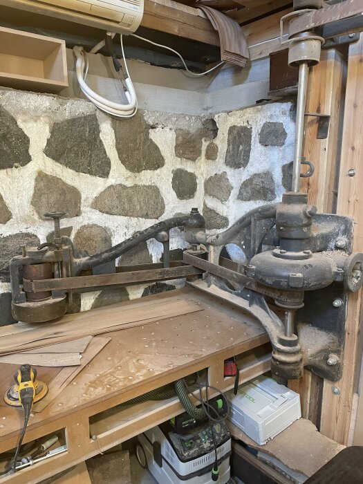 Gamla möbeltappmaskinen i en snickarverkstad med stenmur i bakgrunden och verktyg på arbetsbänken.