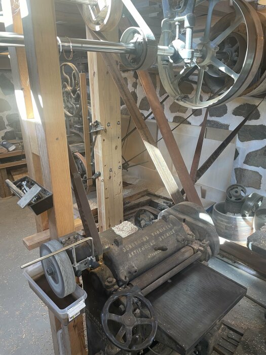 En gammal, mekanisk möbeltappmaskin märkt "Jönköpings Söner" i ett solbelyst snickeri, med stora drivhjul och träråmaterial.
