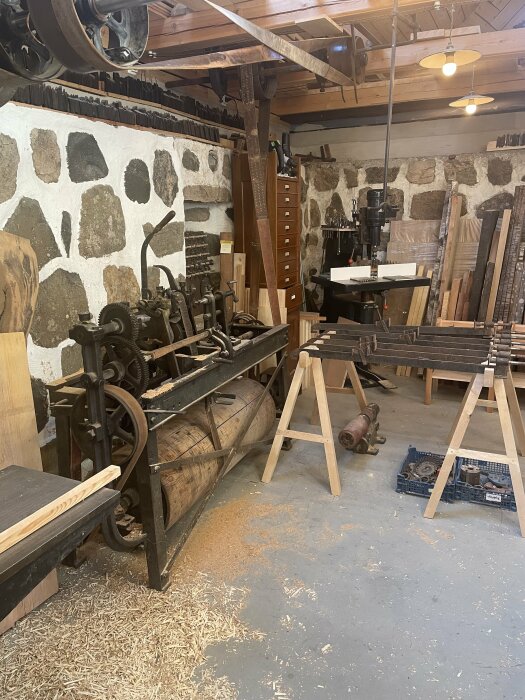 Verkstadsinteriör med äldre träbearbetningsmaskiner och verktyg i ett rum med stenväggar och spån på golvet.