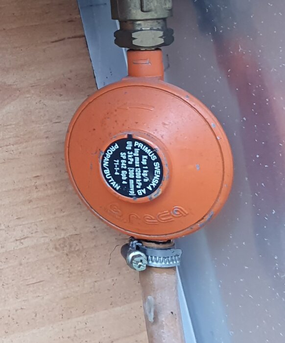 Orange gasolregulator märkt "PRIMUS SVERIGE" fastsatt på en kopparledning vid en husvagn.