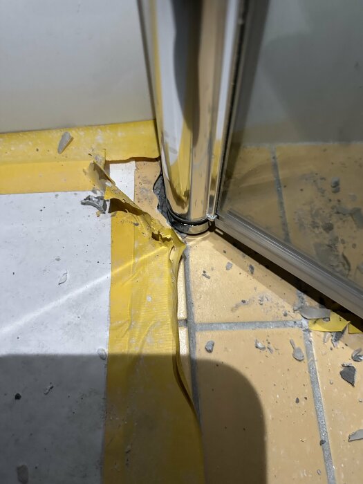 Skadat hörn av gult tätskikt i badrum vid nedsänkning där en duschvägg är installerad, med synligt kakelfix och spill.