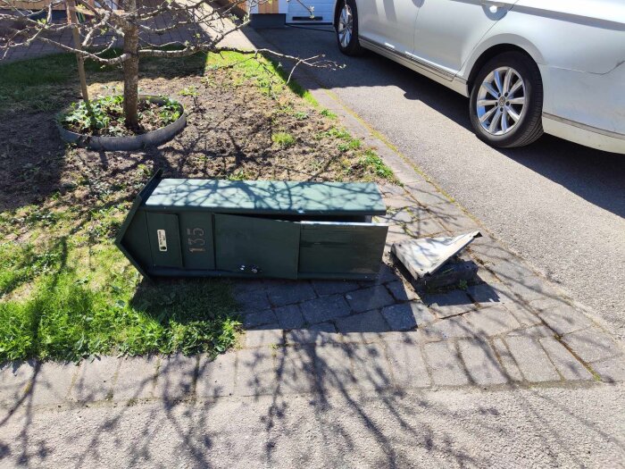 Öppen grön postlåda omkullvält på en trottoar bredvid en parkerad bil och gräsmatta.