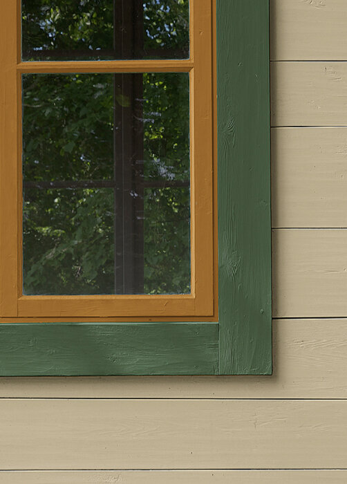 Hörn av en stuga med fasad i vetegrå, Övedsgrön färg på fodret och antikgrön runt ett fönster med orange karm.