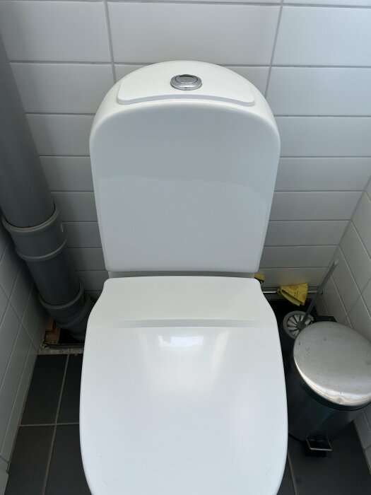 Vit toalettstol i badrum med öppen sitslock, synlig avloppsrör och pedalhink vid sidan.