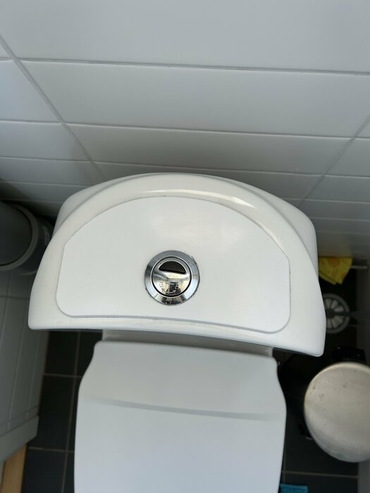 Översikt av en äldre vit toalettstol sett uppifrån, med fokus på spolknappen i behov av reparation.