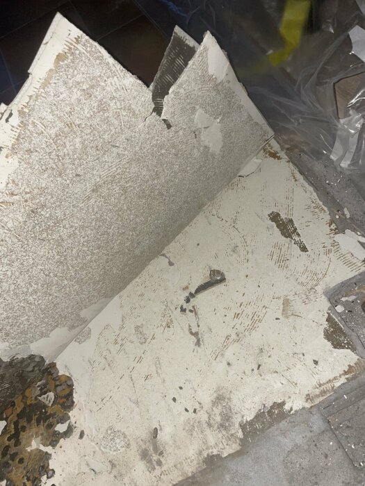 Gammal avrivet golv med synlig underliggande plastmatta och ljusgrått pappmaterial, misstänkt asbest.