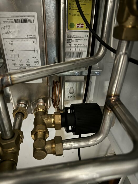 Värmesystem med kopparledningar och en svart pump, nära en vit etikett på en apparat.