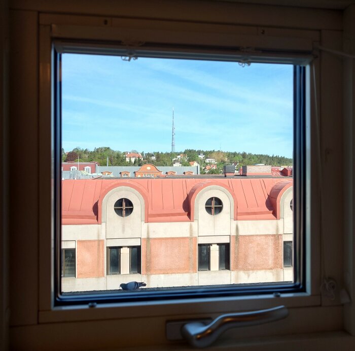 Utsikt genom fönster på stadsbebyggelse med röda tak och träd i bakgrunden under blå himmel.