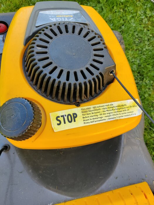 En gul gräsklippare med en svart tändningsspole och instruktionsetikett som anger att man ska fylla på med olja före start.