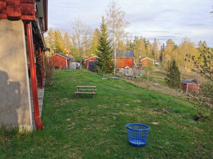 Altan med gräsyta, en bänk och blå korg vid hus med röda uthus i bakgrunden.