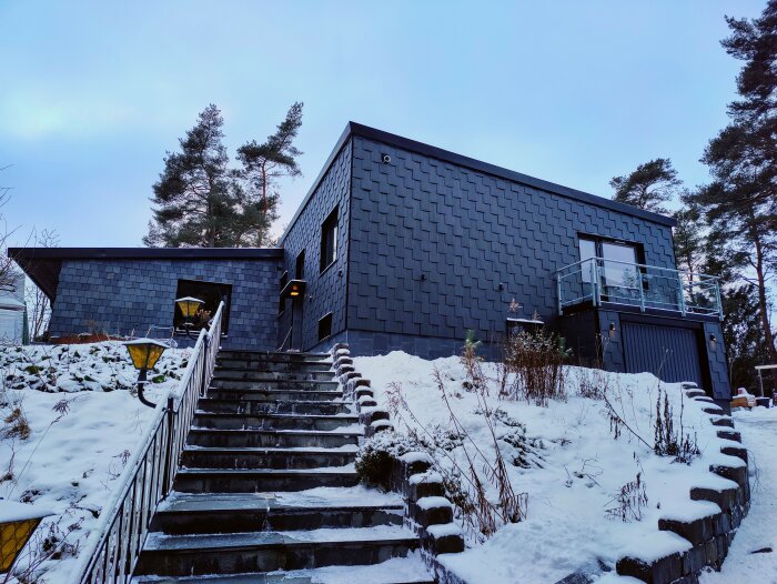 Hus med skiffertak och skifferklädd fasad i vinterlandskap, snö på marken och stentrappa.