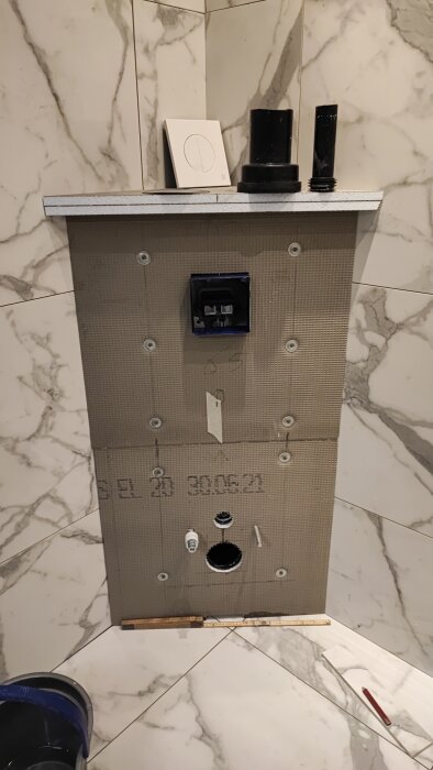 Obehandlad toalett med synlig väggmodul och väntande installering av plattor, med verktyg synligt på golvet.