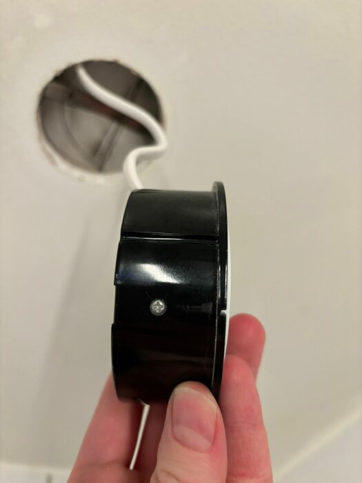 Hand som håller en svart spotlight framför en vit vägg med synligt installationshål.