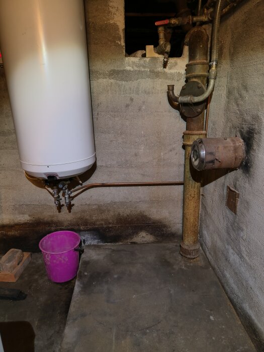 Varmvattenberedare med rör och en rosa hink på betonggolv nära avloppsbrunnen och gjutjärnsrör.