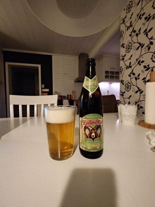 En öppnad flaska tysk maibock och ett glas med guldgul öl på ett vitt köksbord.