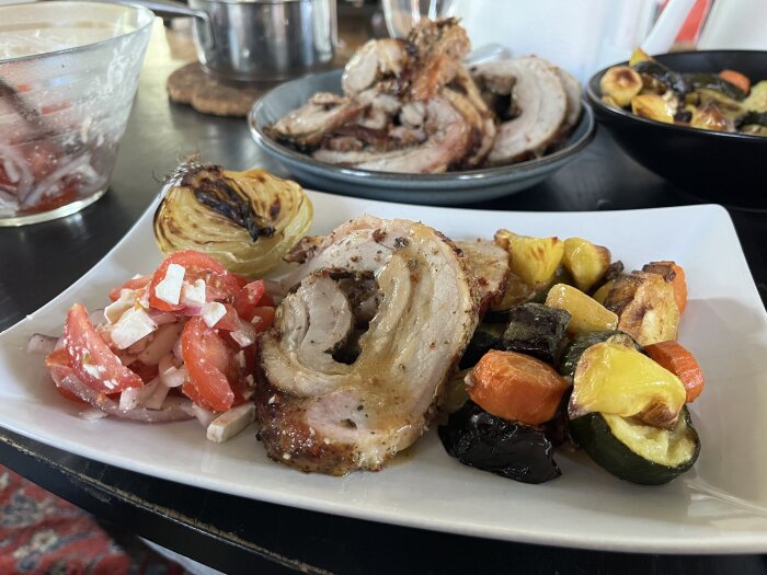 Långstekt porchetta med ugnsstekta grönsaker, grillad lök och tomatsallad med fetaost på en tallrik.