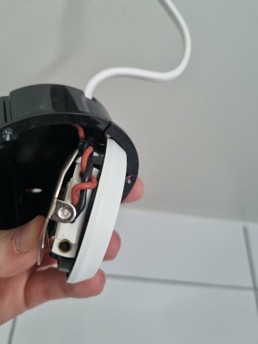Hand håller upp en öppnad elektrisk dosa med synliga sladdar och anslutningar mot en vit vägg.