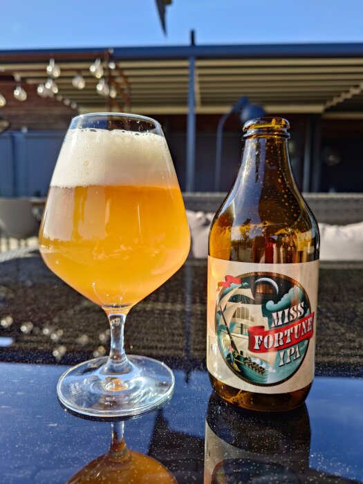 Öppen IPA-flaska och ett glas öl på ett solbelyst utomhusbord.