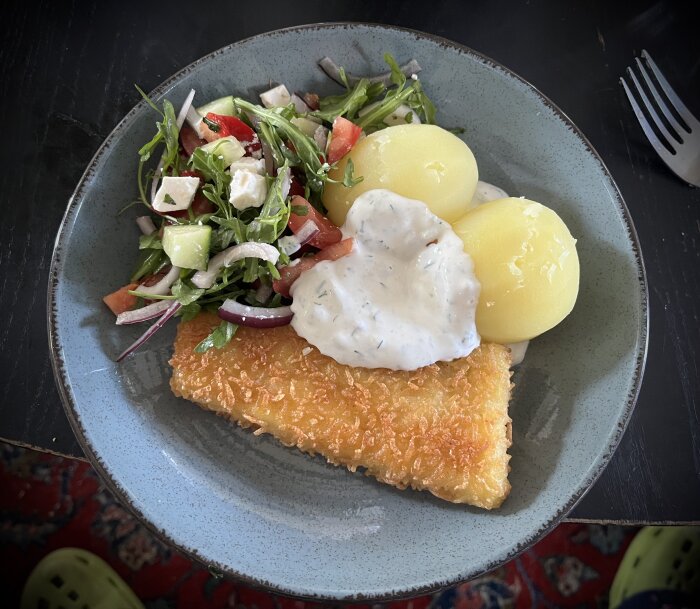 En tallrik med panerad torsk, kokt potatis och fetaostsallad toppad med dill- och citronsås.