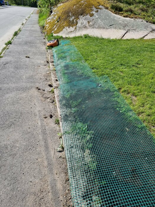 Grön skyddsmatta längs en slänt vid trottoar, en robotgräsklippare fastnar nära kanten.
