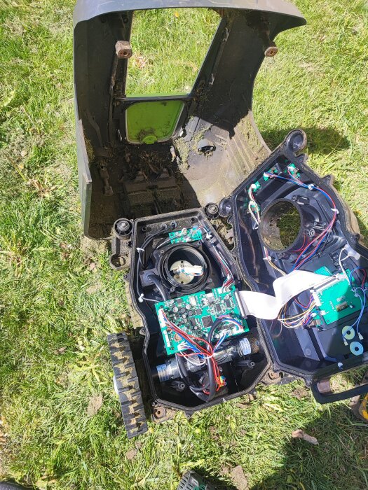 Öppet robotgräsklipparskal på gräs med synliga elektroniska komponenter och batteri under underhåll.