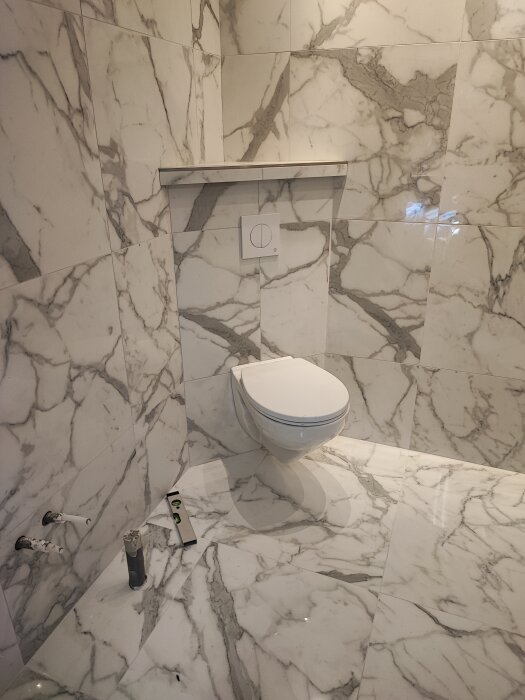 Nyfogat badrum med marmormönstrade kakelplattor och toalett, väntar på spolknappsmontering.