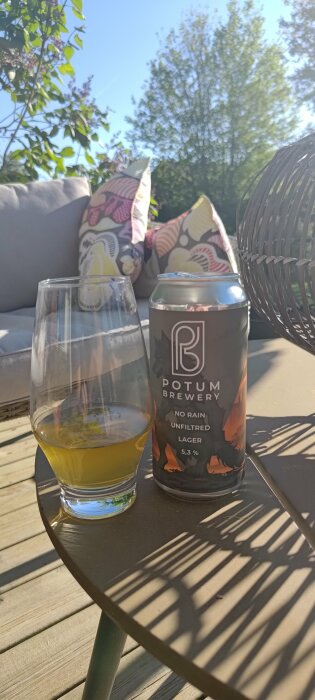 Ett glas öl och en burk från Potum Brewery på ett bord på altan i solsken, omgivna av grönska och kuddar.