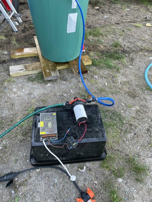 En gammal 12V-pump kopplad till batteri och omvandlare utomhus bredvid en grön behållare.